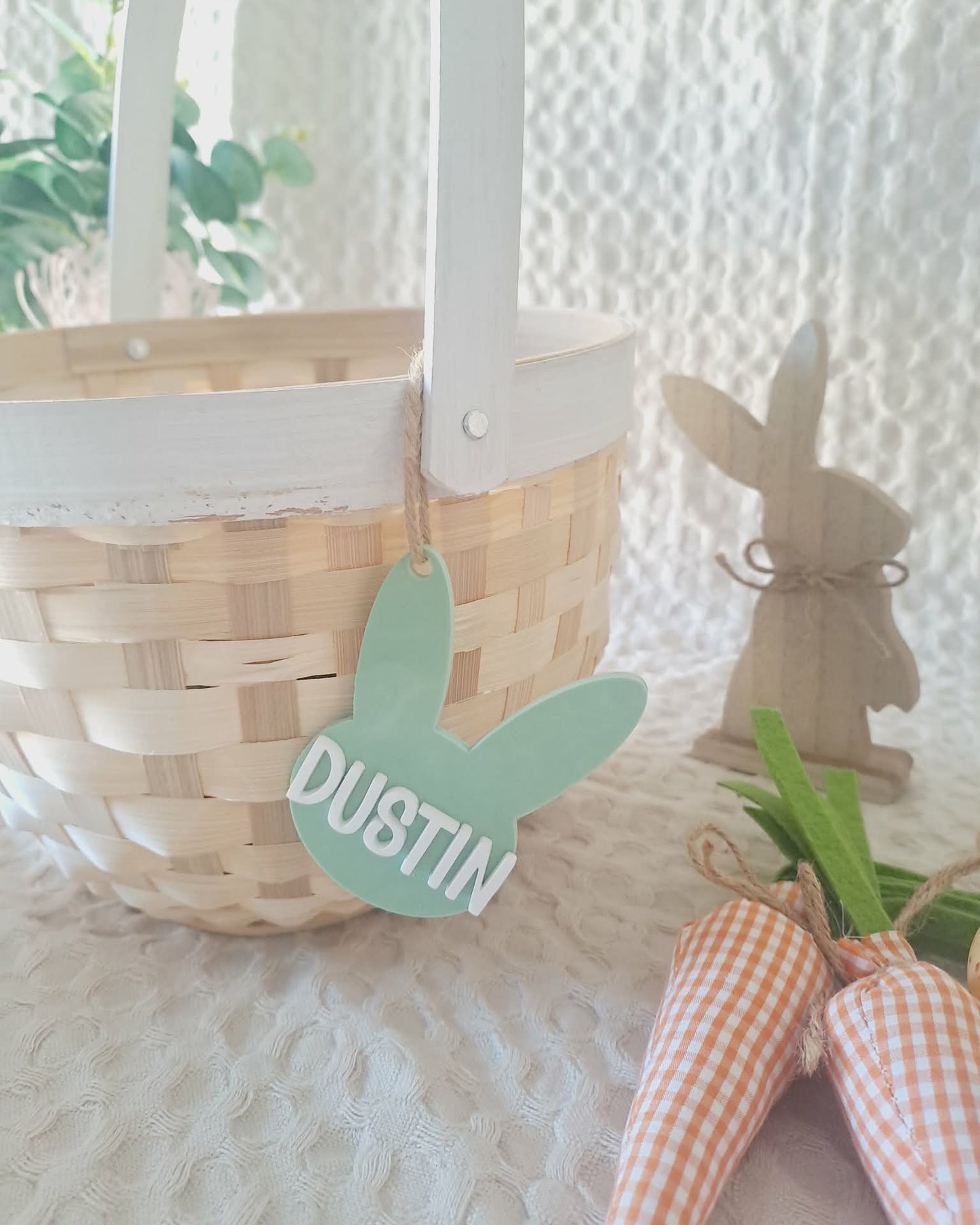 Wicker basket with a green 'Dustin' tag, wooden rabbit figurine, and checkered bow on a textured white surface.