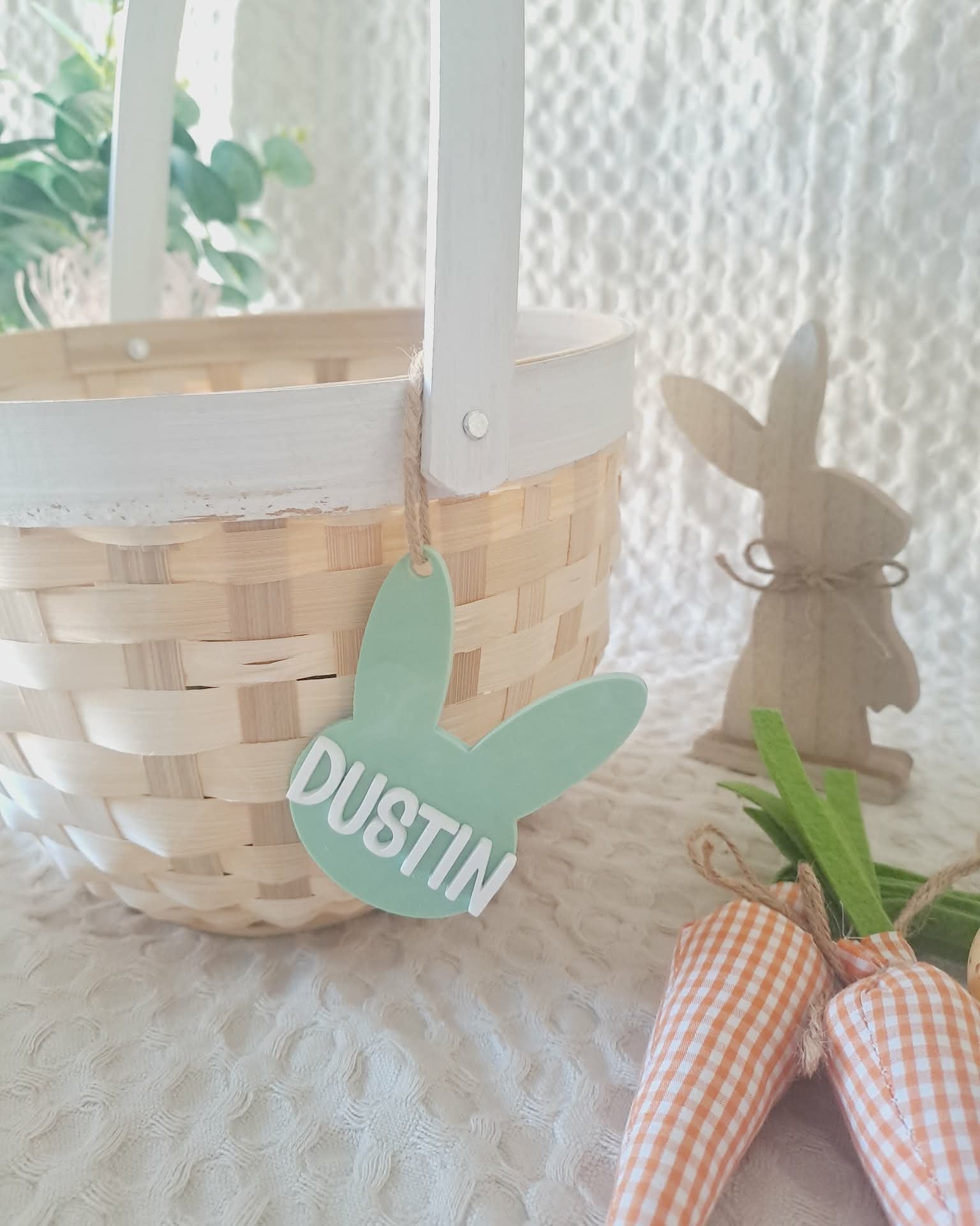 Wicker basket with a green 'Dustin' tag, wooden rabbit figurine, and checkered bow on a textured white surface.