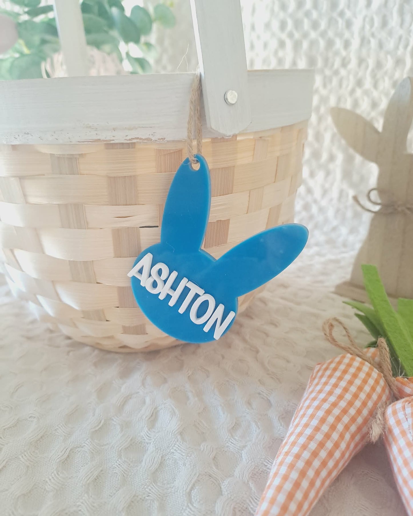 Blue bunny-shaped tag with 'ASHTON' on a woven basket and carrot decoration.