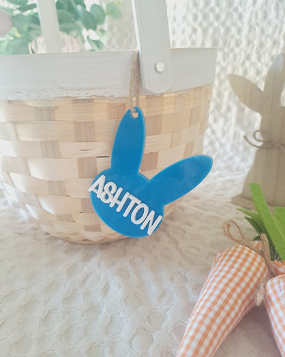 Blue bunny-shaped tag with 'ASHTON' on a woven basket and carrot decoration.