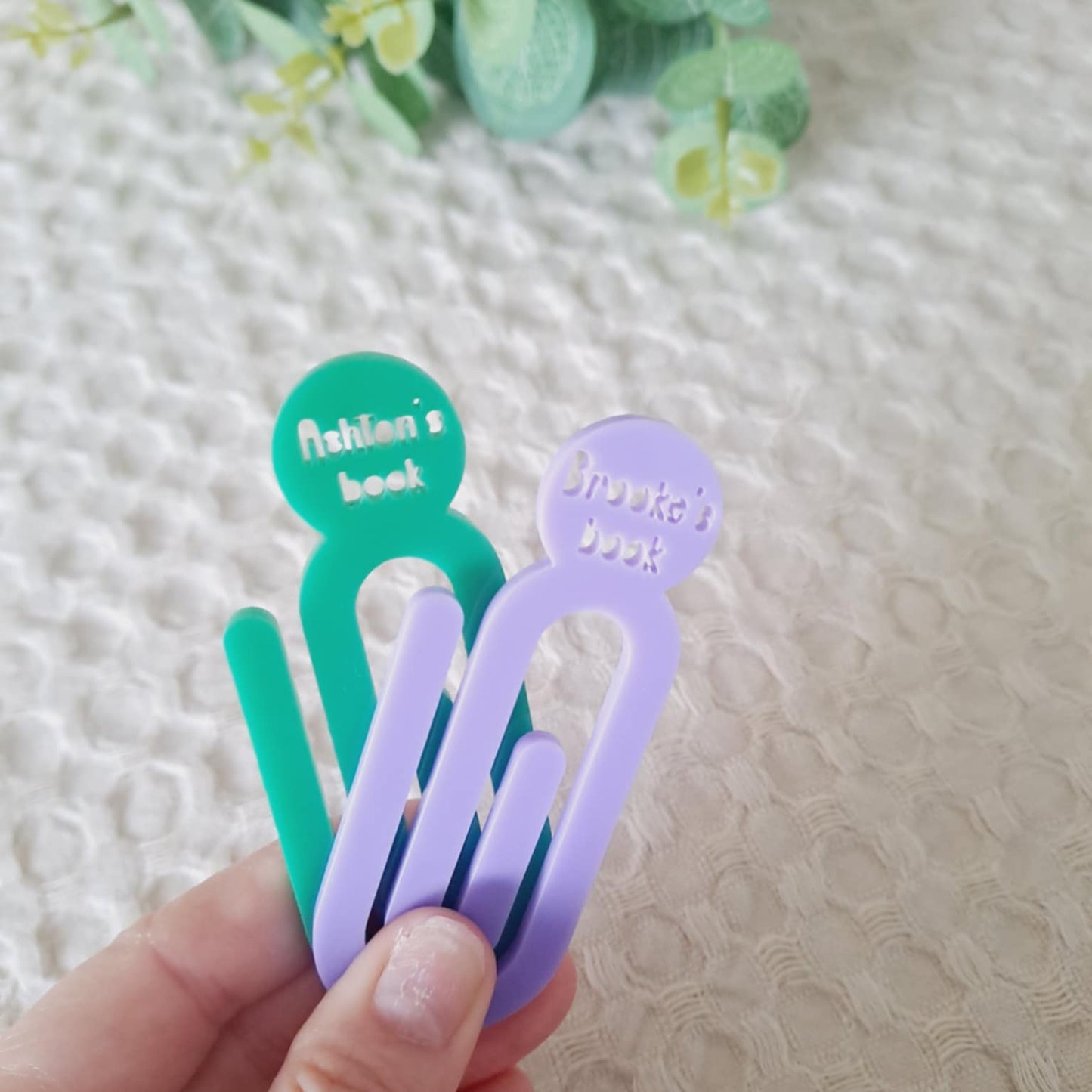 Hand holding two colorful paperclip bookmarks with text on a textured surface.